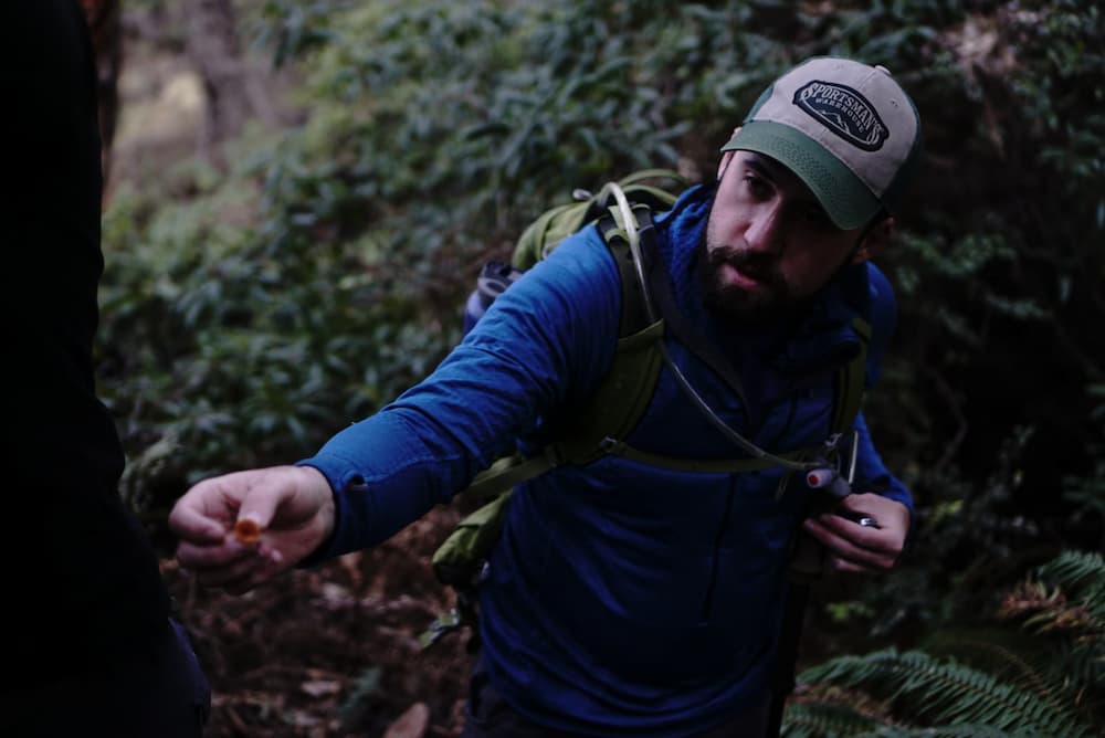 A white man with a beard, wearing a backpack and holding an orange mushroom in the woods